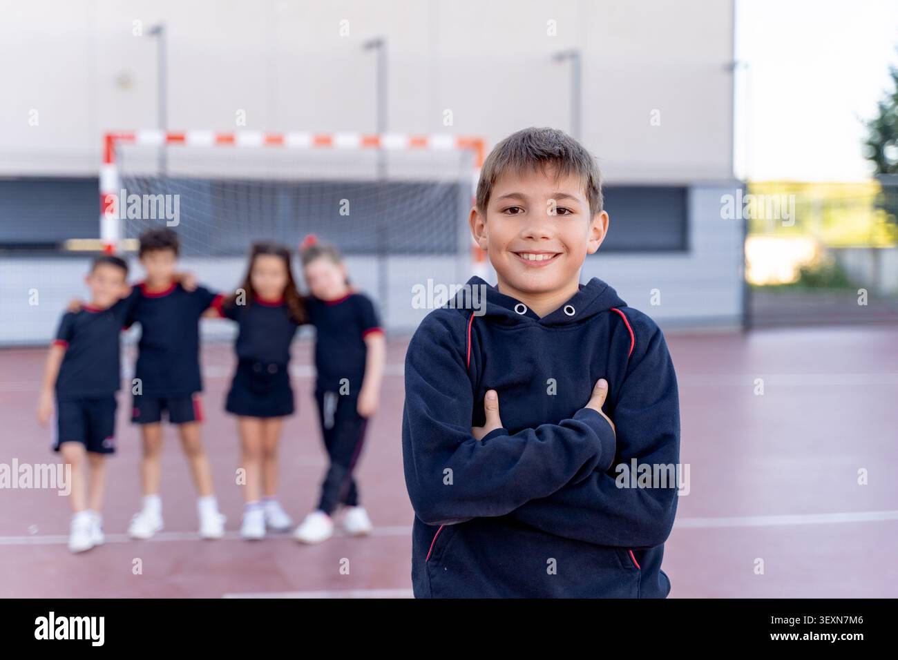 Scolaro felice in piedi con le braccia incrociate, compagni di classe sfocati sullo sfondo su un campo sportivo scolastico con un obiettivo Foto Stock