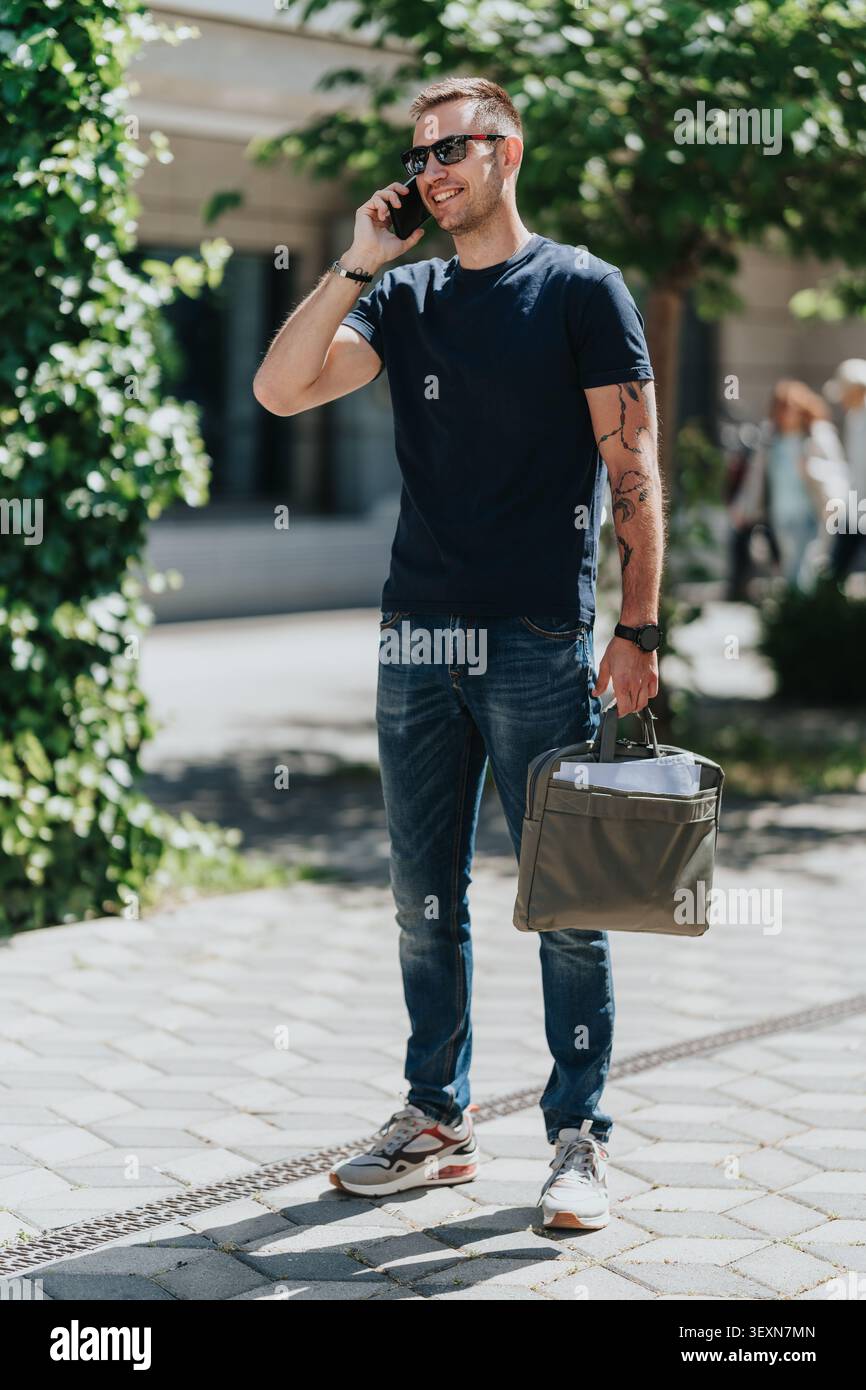 Giovane uomo d'affari sorridente che parla al telefono mentre porta con sé una borsa portadocumenti all'aperto sul marciapiede della città Foto Stock