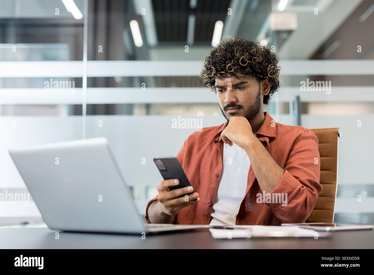 Giovane indiano dai capelli ricci che si concentra con attenzione sul suo smartphone mentre lavora a una scrivania con un computer portatile in un ufficio moderno, sentendosi attento e concentrato Foto Stock