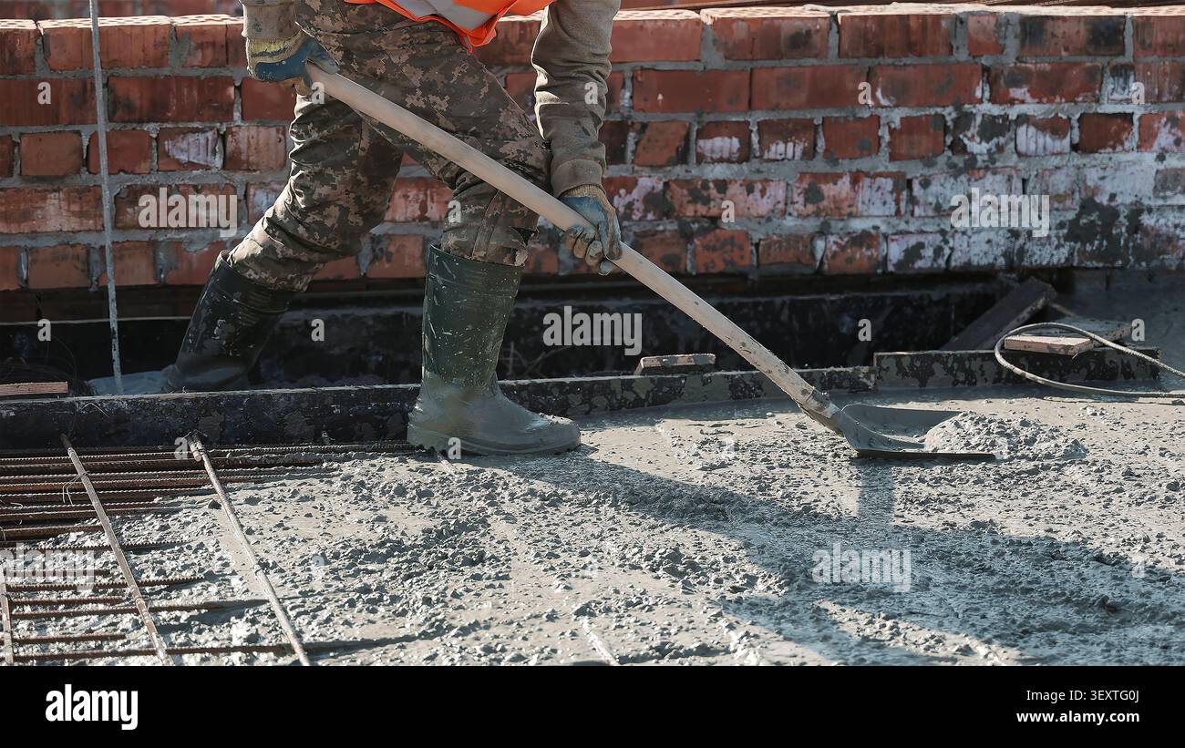 Operaio che spande calcestruzzo bagnato nel cantiere. L'operaio edile corto in stivali e guanti diffonde calcestruzzo umido con una pala con manico lungo Foto Stock