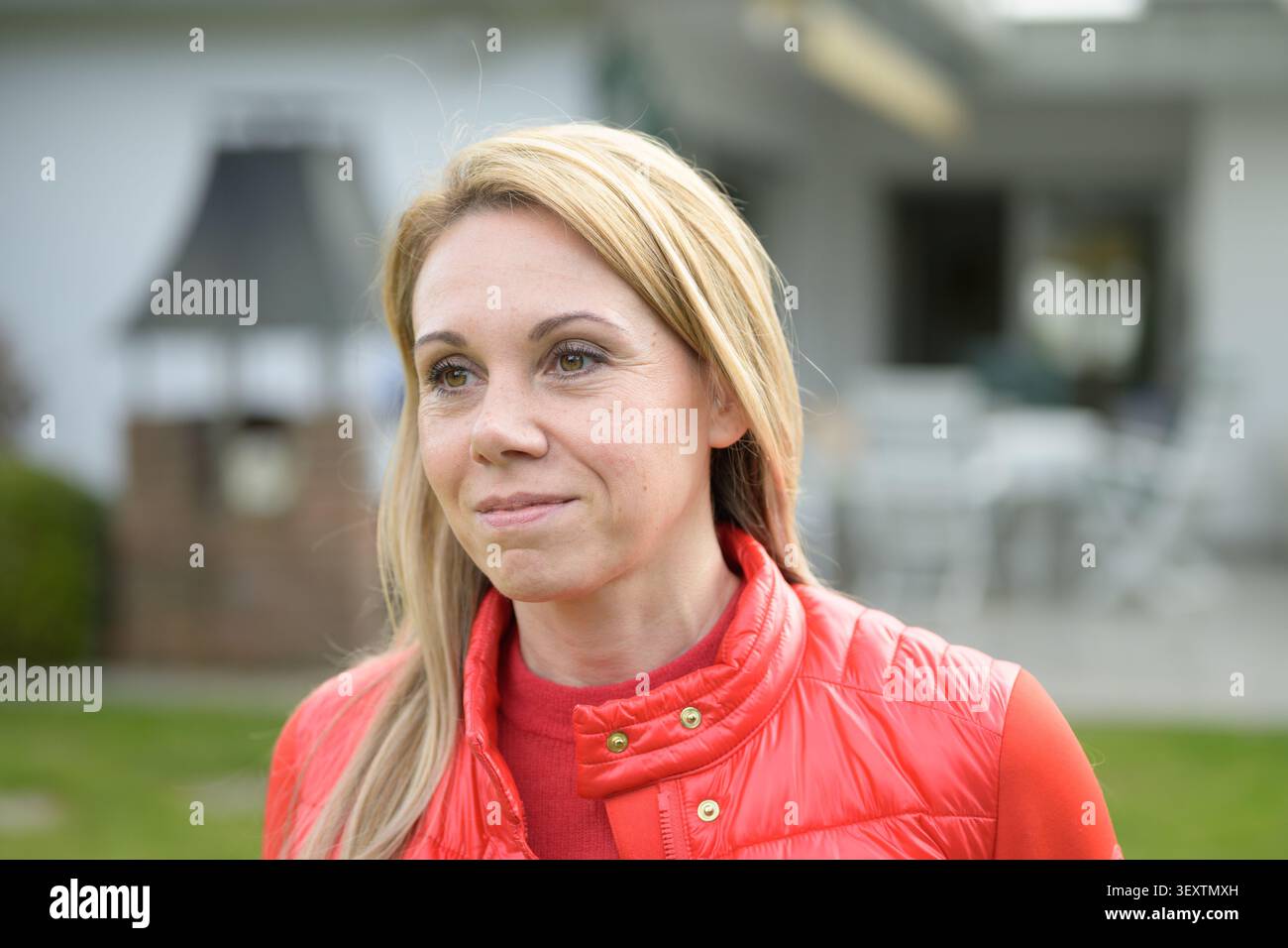 Ritratto all'aperto di una donna bionda che indossa un gilet trapuntato rosso brillante mentre guarda in lontananza in un moderno ambiente residenziale. Foto Stock