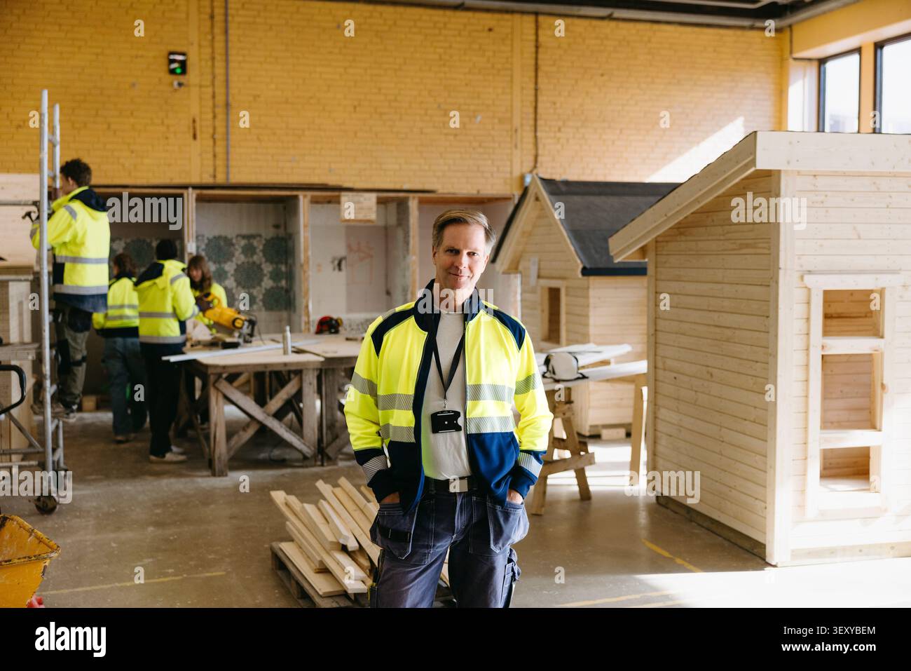 Ritratto sorridente di un istruttore maschio maturo in piedi con le mani in tasca presso l'officina edile Foto Stock