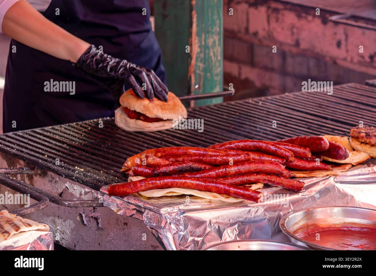 Uomo che prepara un hamburger in un mercato di campagna e grigia salsicce rosse brillanti su una griglia piatta Foto Stock