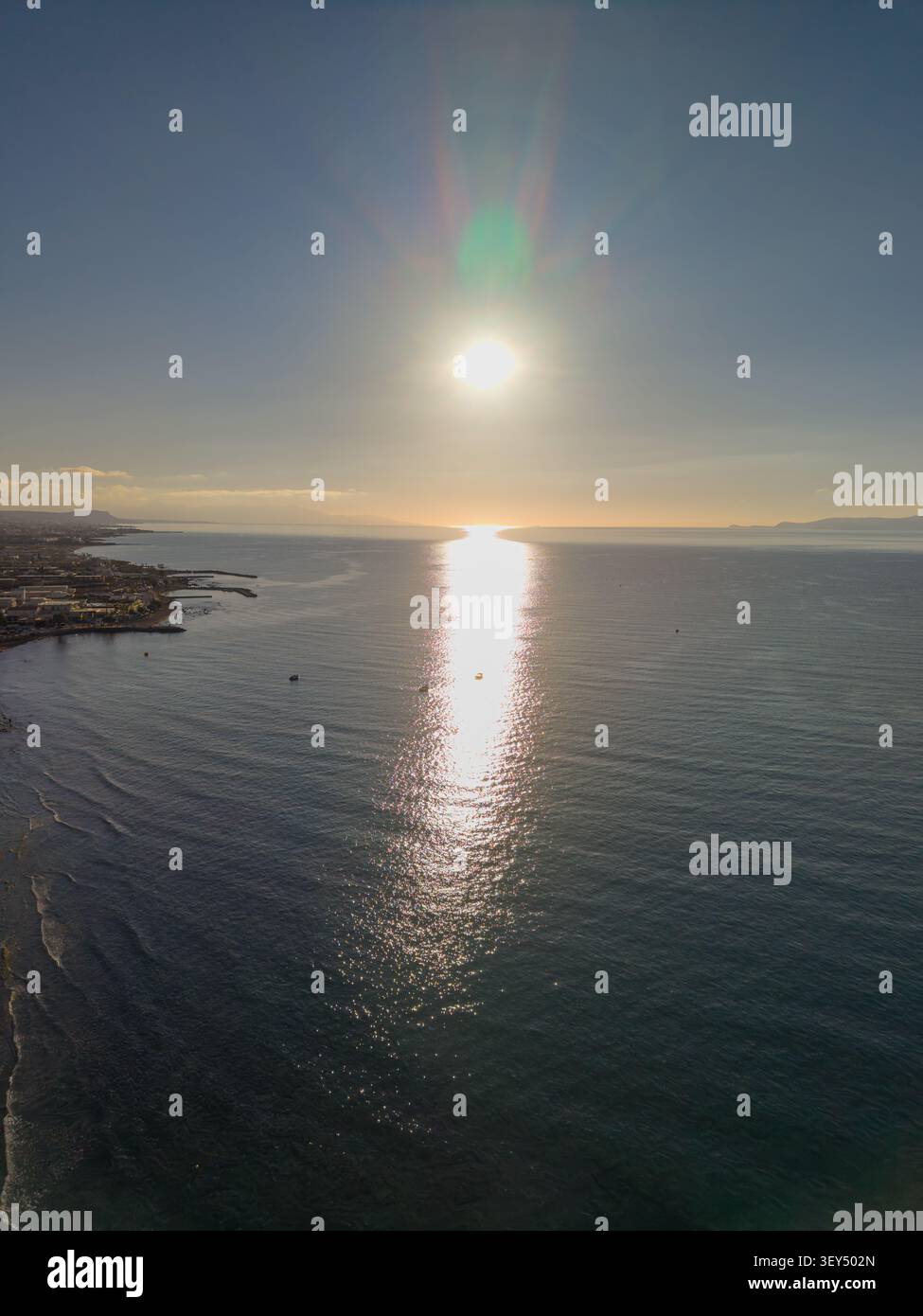 La prospettiva di un drone ad alto angolo cattura un sole luminoso che tramonta sulla superficie del mare creando un forte riflesso di luce dorata vicino a una costa. Foto Stock