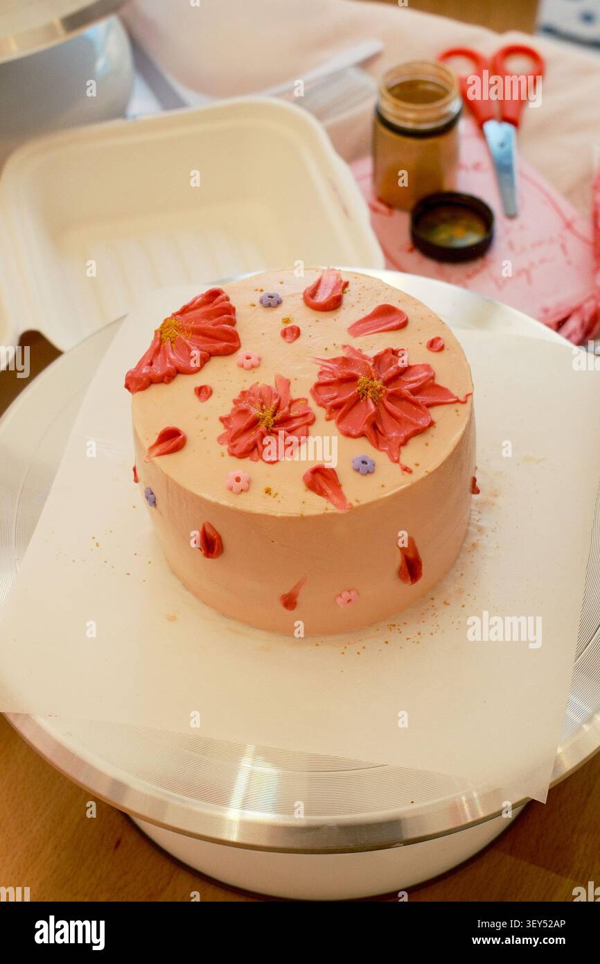 Torta rosa in stile bento con fiori a margherita di crema di corallo, piccoli fiori fondenti e polvere dorata su un tavolo girevole per torte d'argento durante un lavoro di cottura Foto Stock