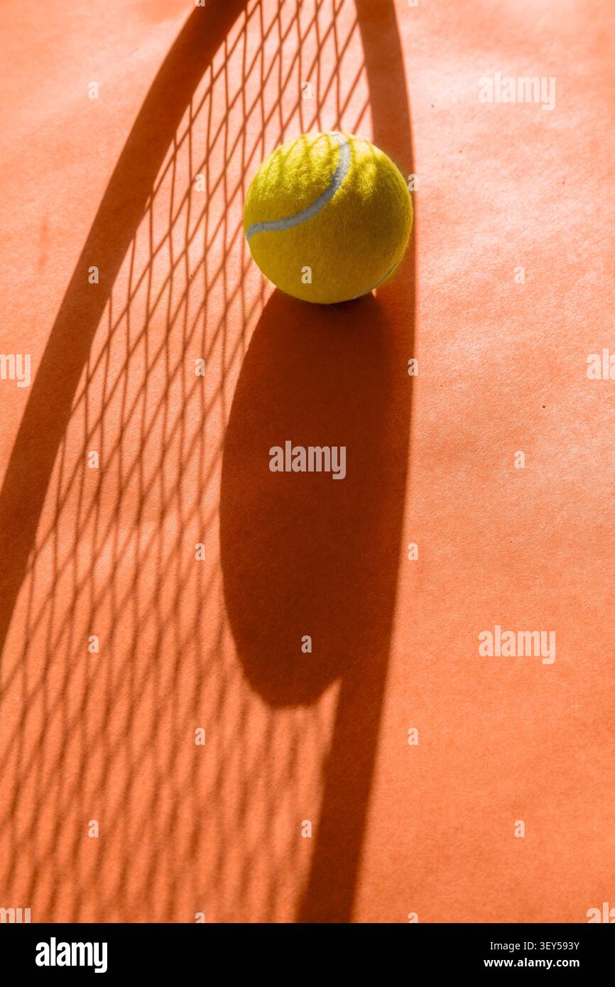 Palla da tennis gialla appoggiata su un campo di argilla arancione con una spettacolare ombra di una racchetta da tennis che crea motivi geometrici. La luce solare intensa enfatizza l'effetto tex Foto Stock