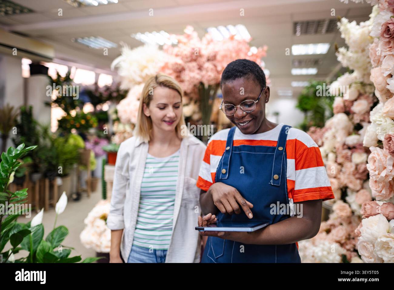 Fiorista che mostra le composizioni floreali su un tablet a un cliente in un negozio di fiori, piccole imprese e concetto tecnologico Foto Stock