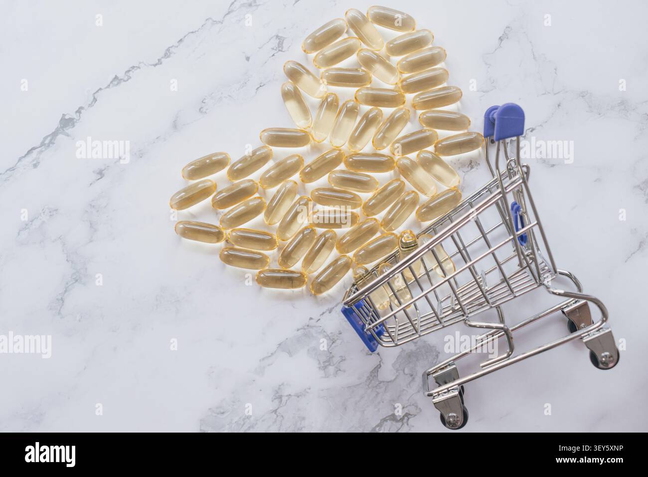 Colpo orizzontale ad angolo alto di capsule di olio di pesce omega dorato disposte a forma di cuore con un piccolo carrello su fondo di marmo bianco sotto B. Foto Stock