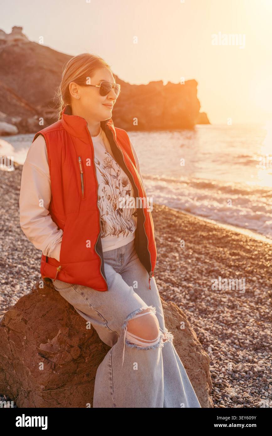 La spiaggia al tramonto della donna si rilassa su una spiaggia rocciosa indossando un gilet rosso e occhiali da sole durante l'ora d'oro al mare Foto Stock