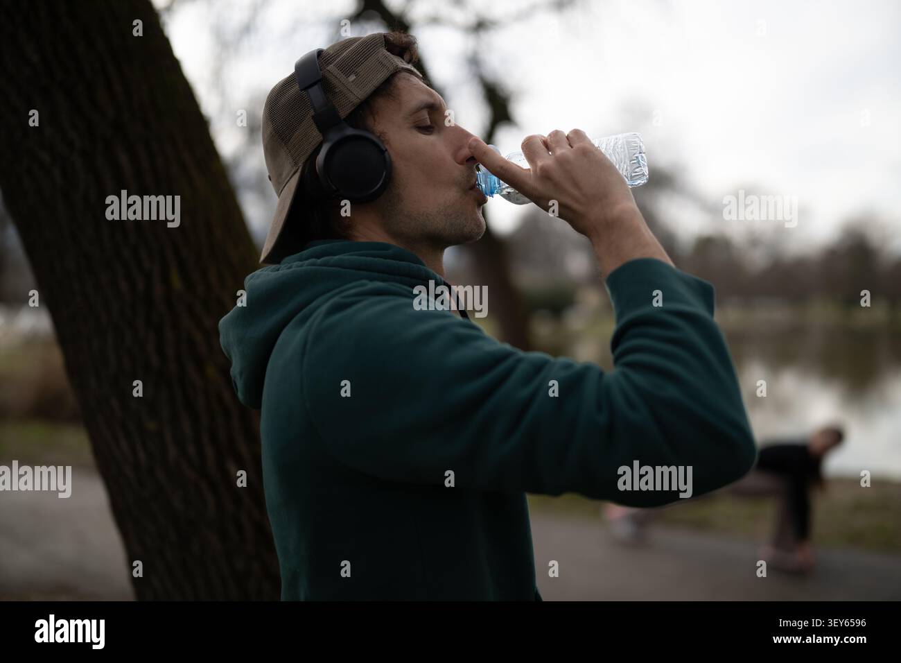 Il giovane beve acqua mentre indossa le cuffie e un berretto in un parco Foto Stock