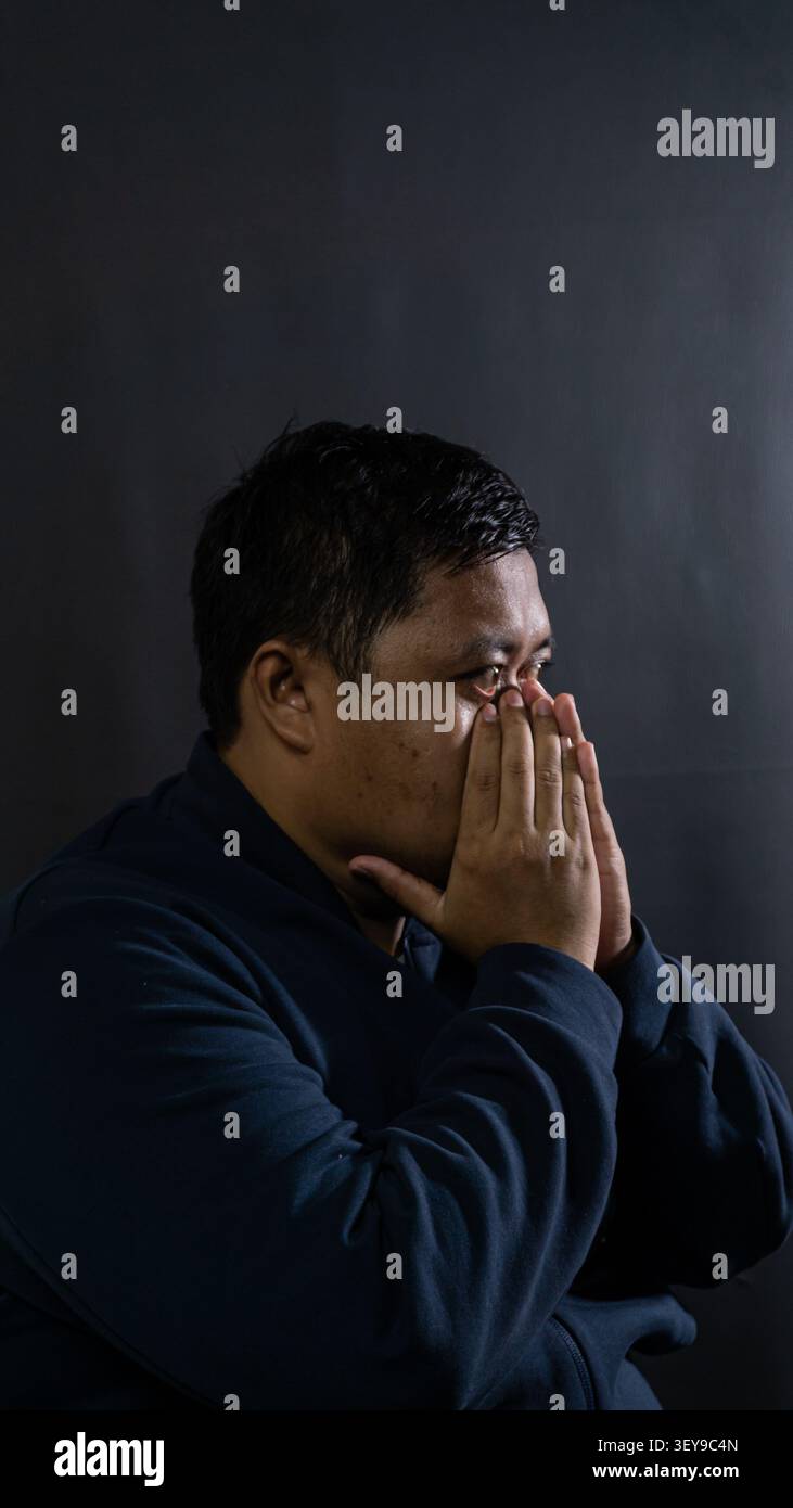 Un profilo laterale ravvicinato di un maschio indonesiano ansioso che stringe entrambe le mani davanti alla bocca mostra profonda preoccupazione, lotta interna e alto st Foto Stock