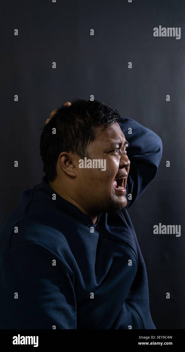 Un maschio indonesiano emette un urlo a bocca aperta di rabbia intensa e profonda frustrazione mentre gli si afferra la schiena della testa con entrambe le mani Foto Stock