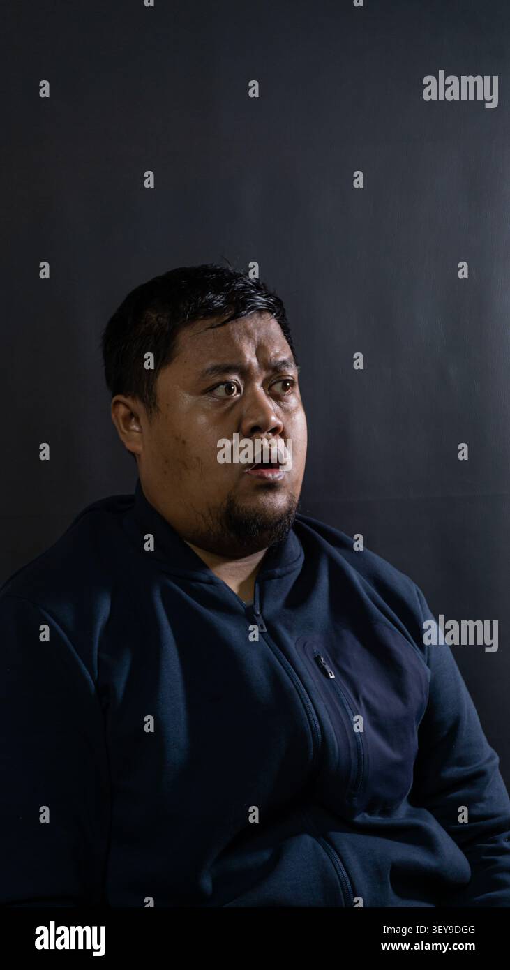 Un adulto indonesiano sconvolto e incredulo, la sua bocca si agita leggermente, su un profondo sfondo grigio scuro in studio Foto Stock