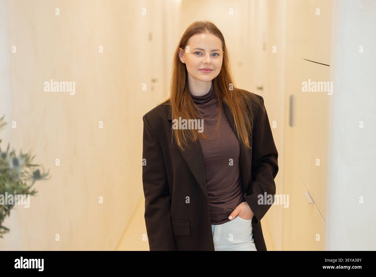 Giovane donna d'affari sicura di sé con un elegante blazer marrone che sorride in un moderno e luminoso corridoio d'ufficio Foto Stock