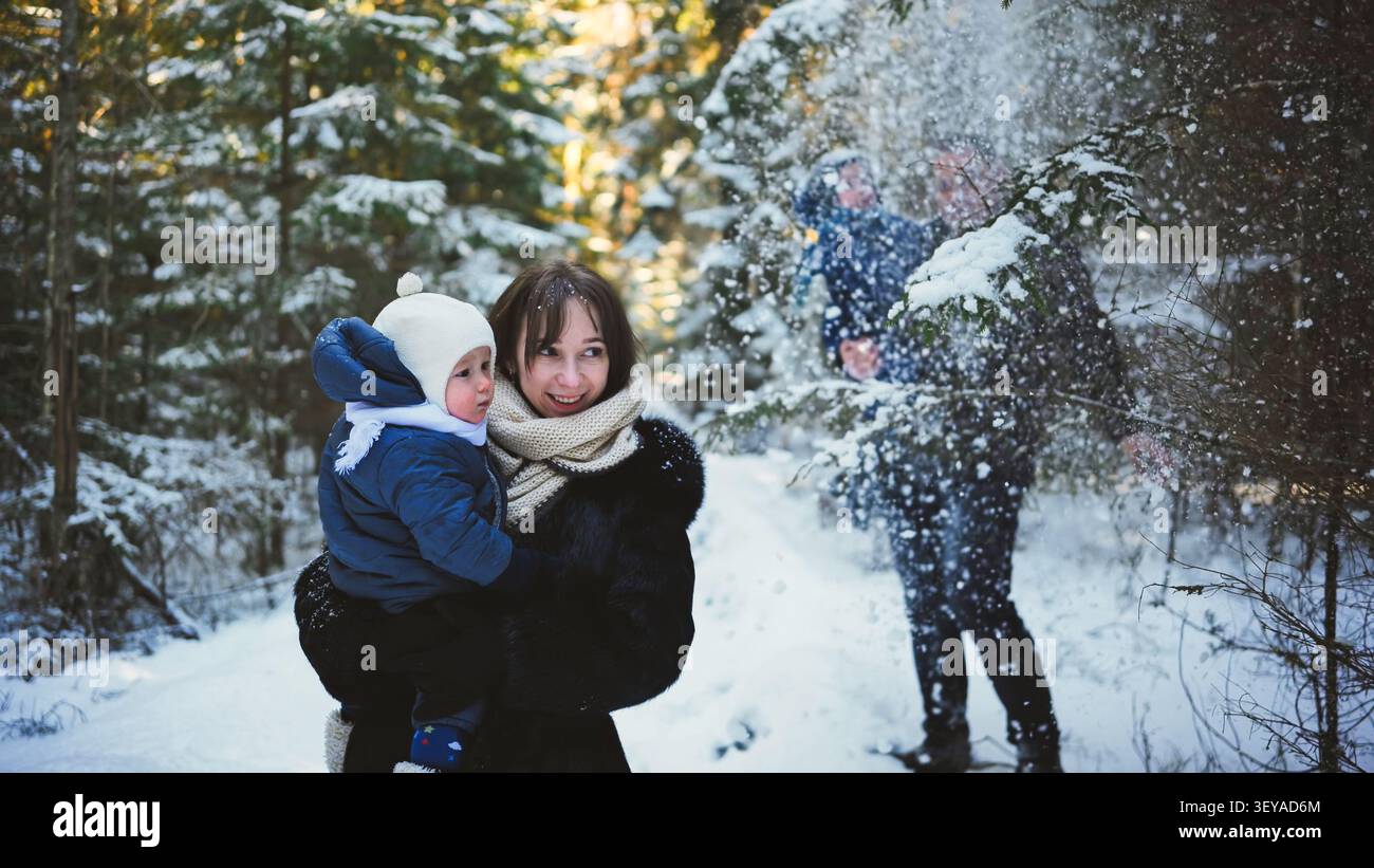 Foresta invernale con i genitori che giocano, la madre abbraccia il bambino mentre il padre pulisce i rami di pino carichi di neve Foto Stock