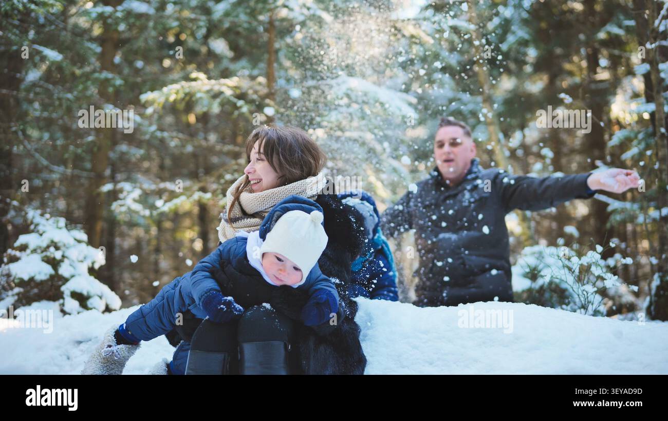 Famiglia felice che ride mentre gioca nella foresta innevata, circondata da alberi carichi di gelo e fiocchi di neve alla deriva Foto Stock