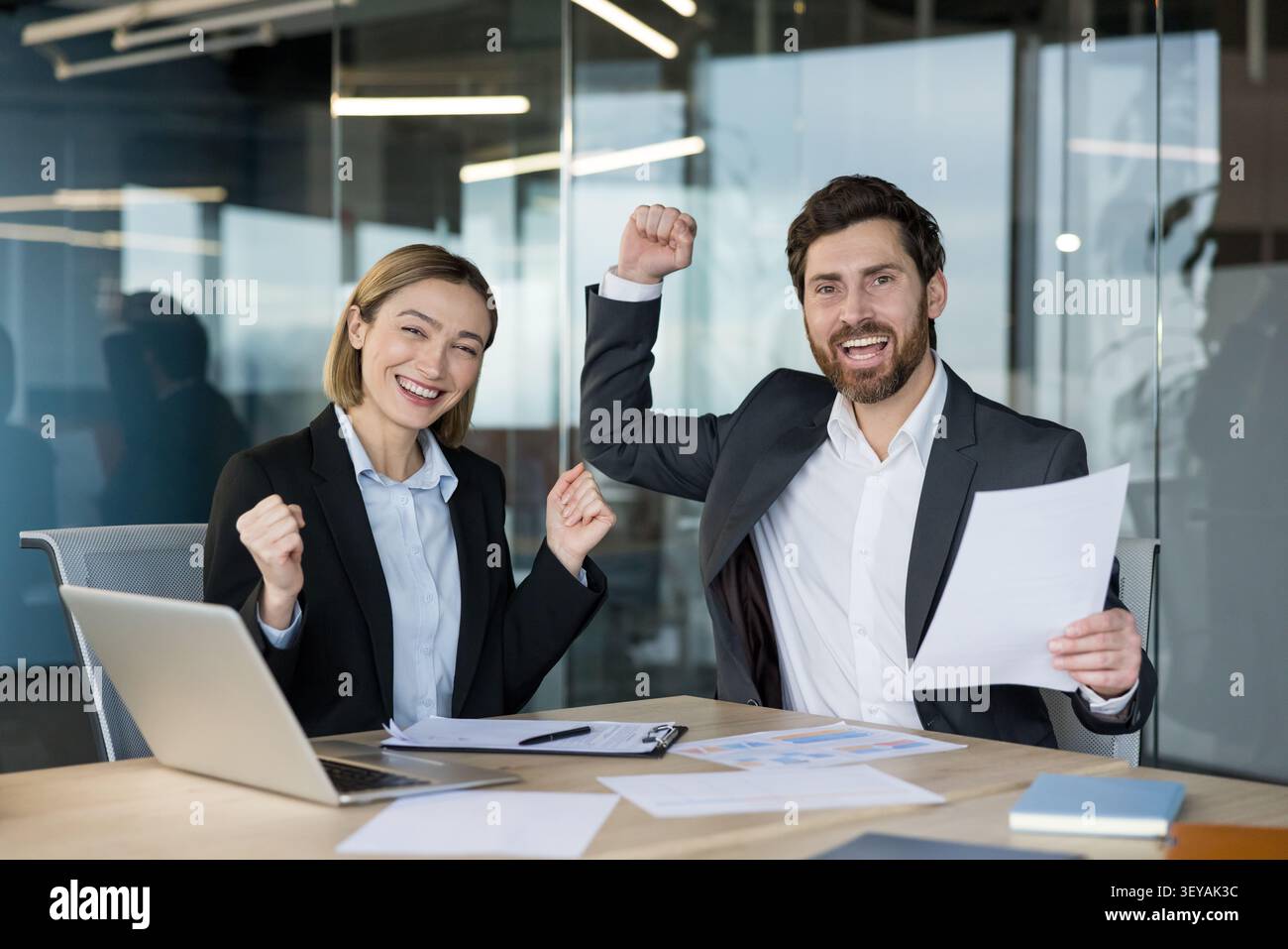 I colleghi d'affari che indossano abiti formali sono seduti a una scrivania e festeggiano felicemente il successo del lavoro di squadra con pugni alzati e sorrisi travolgenti in un ambiente d'ufficio moderno Foto Stock