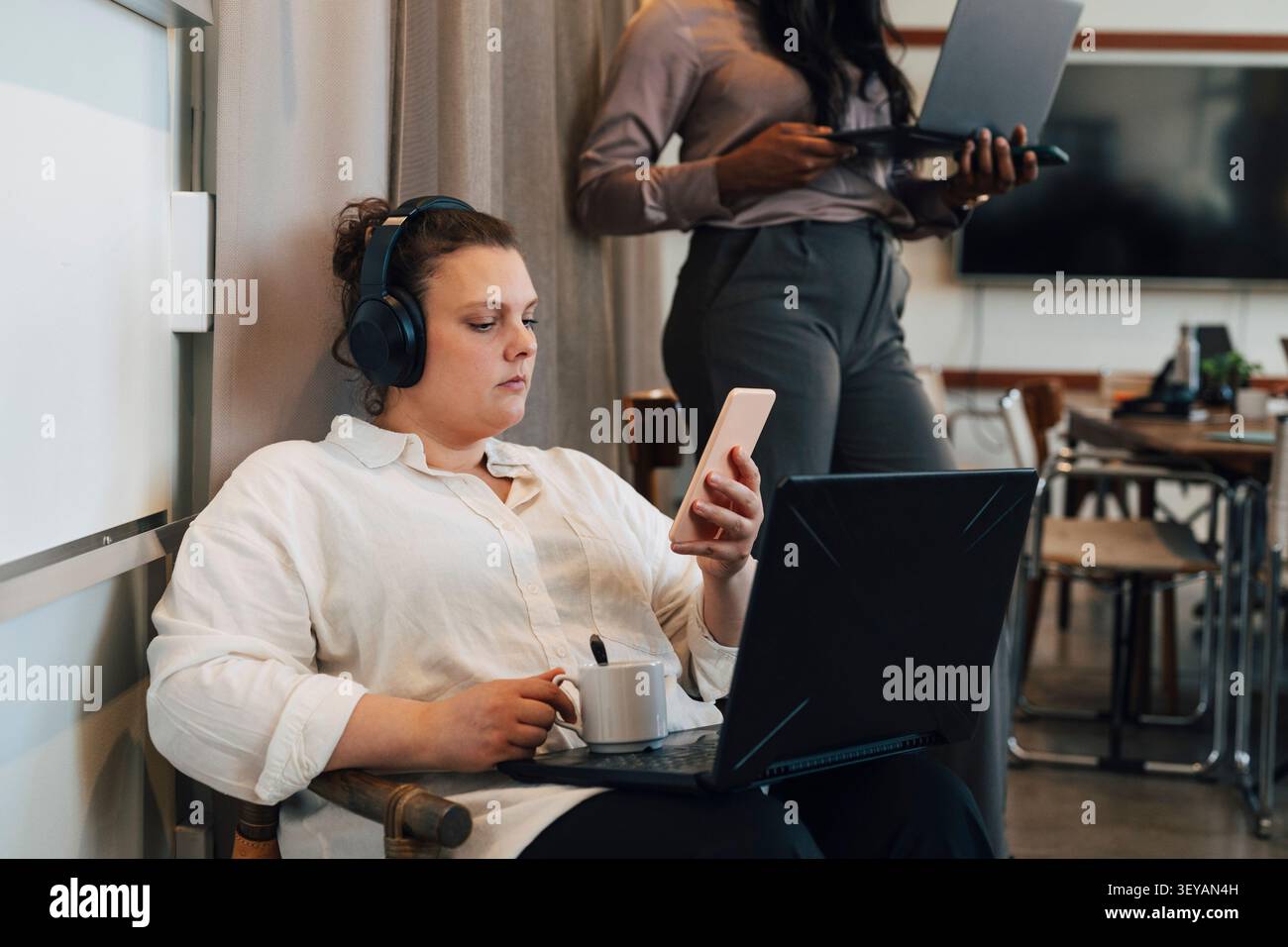Lavoratrice freelance che utilizza il suo smartphone con un laptop aperto in grembo nel moderno spazio di coworking Foto Stock