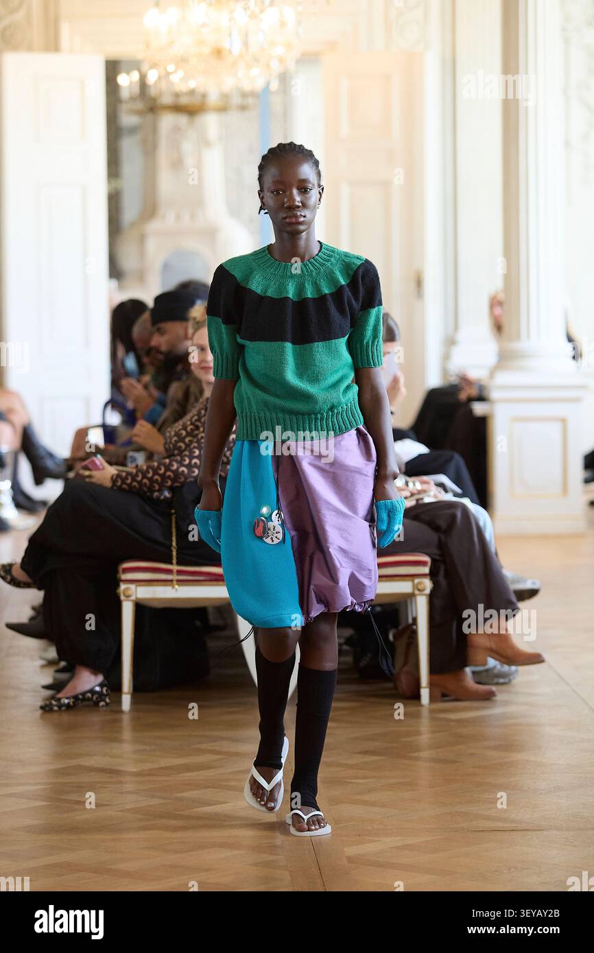 Berlino, Germania, 2 luglio 2026. Un modello cammina sulla passerella presentando una creazione della collezione Primavera/Estate 2027 (SS27) del designer ANDREJ GRONAU. L'outfit presenta un pullover a maniche corte in maglia verde con un'ampia striscia nera orizzontale sul petto, abbinato a una gonna asimmetrica multitesturizzata in maglia blu e tessuto viola drappeggiato ornato da bottoni a spilla. Questo look dallo stile eclettico è rifinito con guanti senza dita blu, calze nere al ginocchio e sandali bianchi con perizoma minimalista. Crediti: Felix Wolf/Alamy Live News Foto Stock