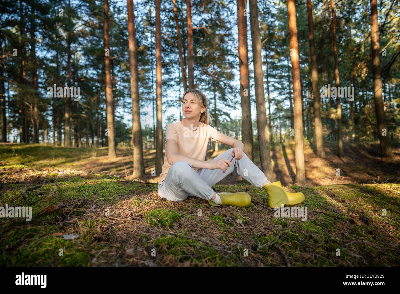 Donna sorridente e tranquilla che si rilassa nella foresta autunnale da sola, seduta sul prato sotto la calda luce del sole. Foto Stock
