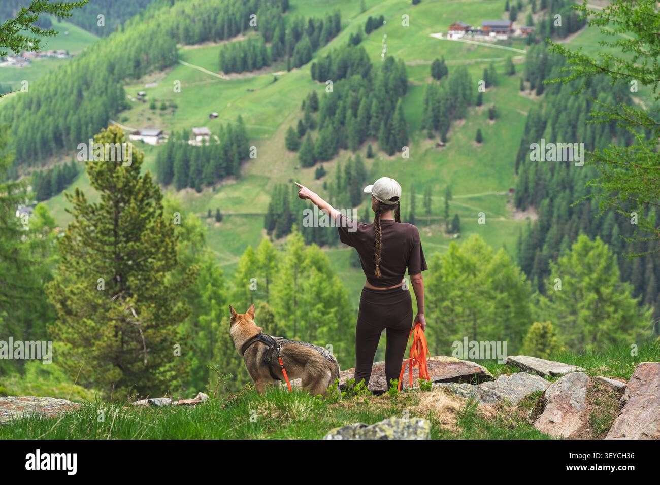 Donna con cane che punta al paesaggio, in un paesaggio montuoso, con donna, cane, cappello, guinzaglio, con erba, alberi, consistenza rocciosa, da un perspecti sopra la testa Foto Stock