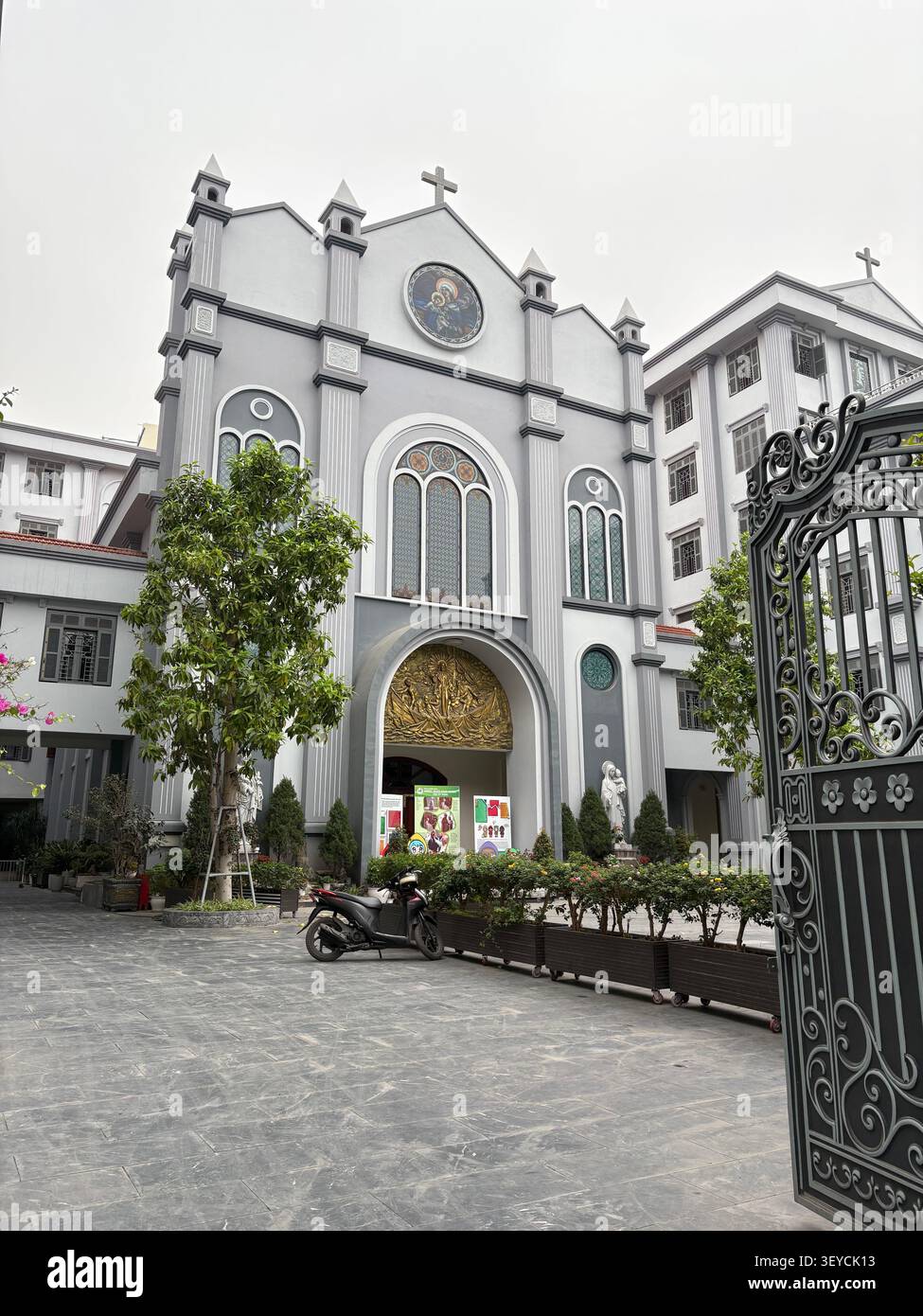 Facciata bianca della chiesa cattolica con finestre ornate in vetro colorato, scultura dorata a rilievo e cancello decorativo in ferro in Vietnam. Architetto religioso urbano - Immagine stock catturata con smartphone