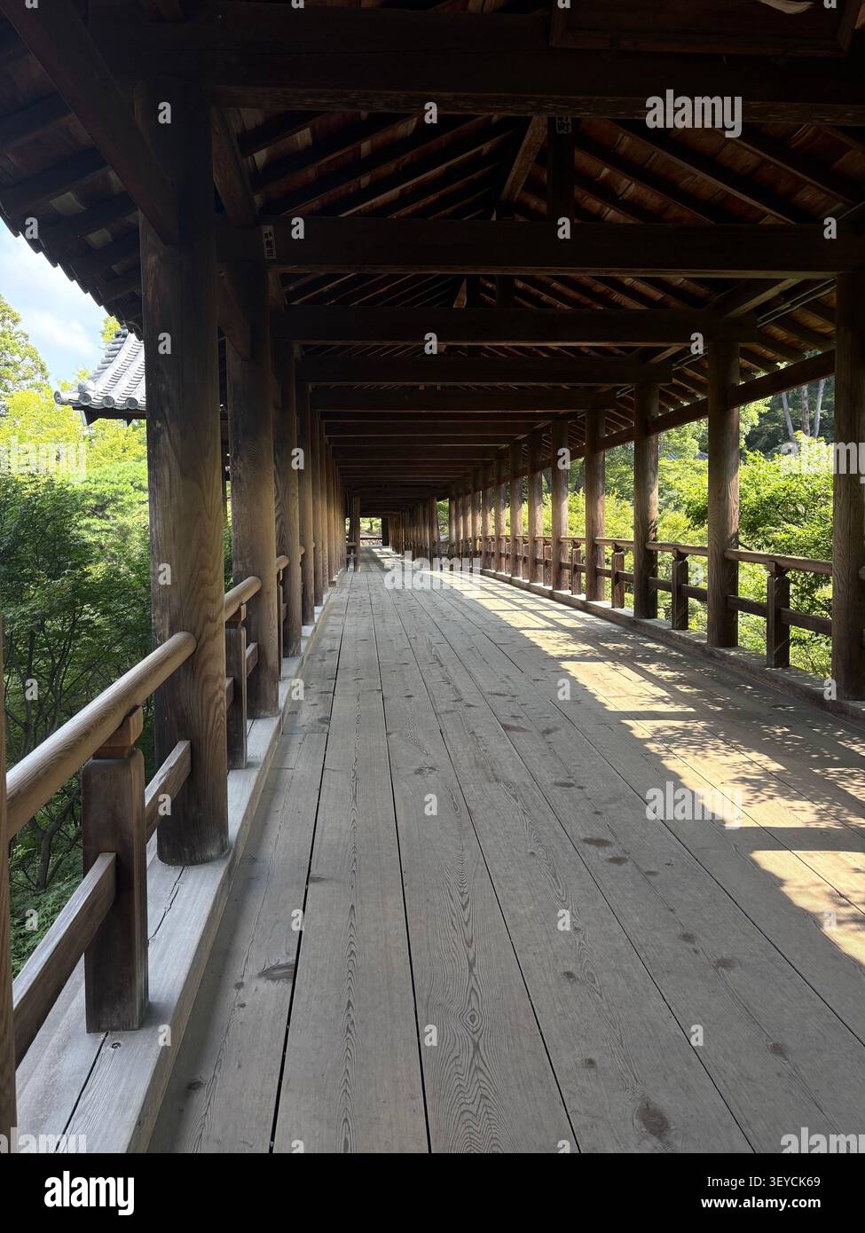 Prospettiva ad angolo basso lungo un tradizionale corridoio in legno presso un santuario shintoista giapponese, con luce solare attenuata che filtra attraverso la struttura a tetto aperto - Immagine stock catturata con smartphone