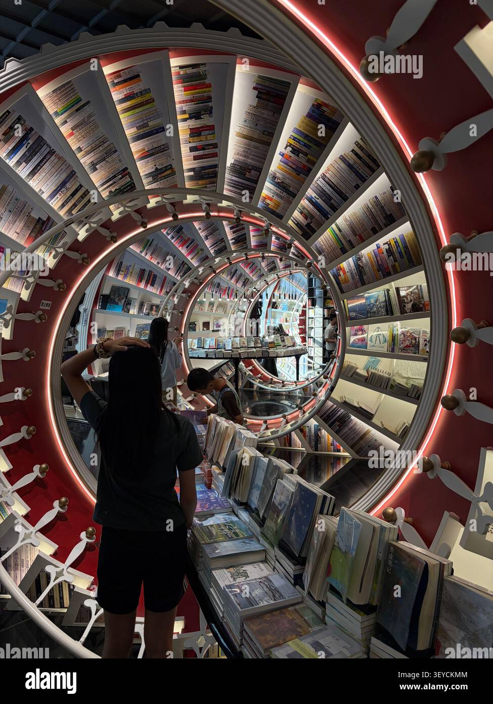 Splendida libreria interna a spirale con ripiani circolari rivestiti con libri colorati, illuminazione al neon rossa e decorazioni bianche ornamentali. Design asiatico contemporaneo - Immagine stock catturata con smartphone