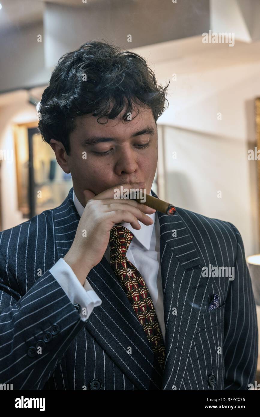Ritratto di un giovane vestito con un abito scuro e appuntito, che fuma un sigaro di lusso. Foto Stock
