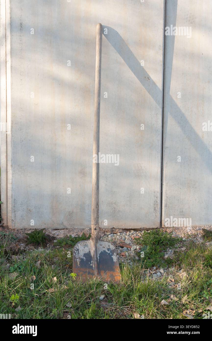 Una pala solitaria poggia su una parete in cemento armato in una giornata di sole. Il contrasto tra la superficie in calcestruzzo ruvido e l'utensile metallico Foto Stock