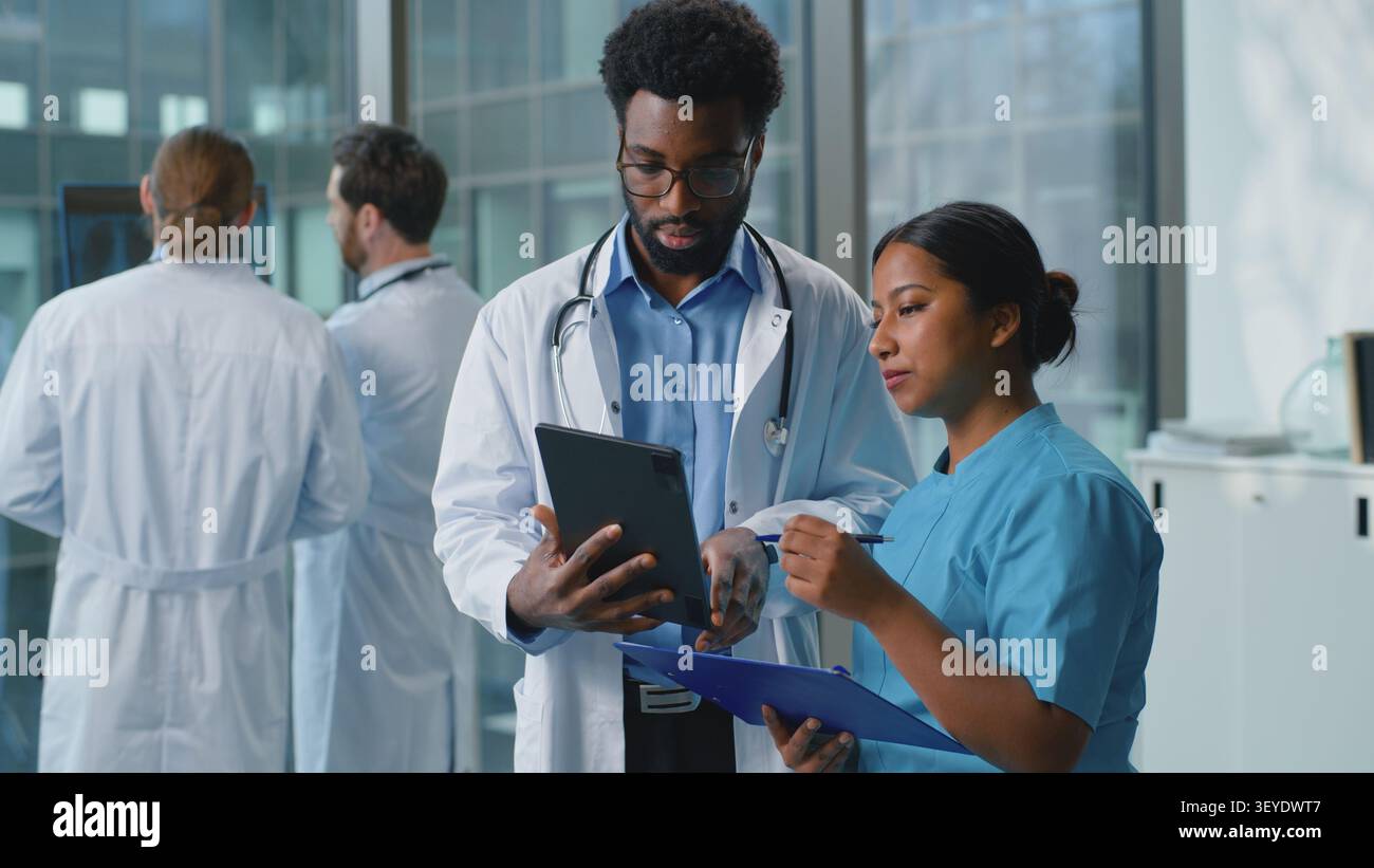 Due medici multirazziali che discutono la diagnosi guardando in tablet digitale in clinica. Uomo afroamericano medico con donna araba indiana infermiera che parla Foto Stock