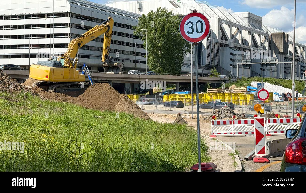 19.05.2026 - Berlino, Germania. Escavatore giallo che scava terra nel cantiere stradale, segnale stradale che indica il limite di velocità in ambiente urbano Foto Stock