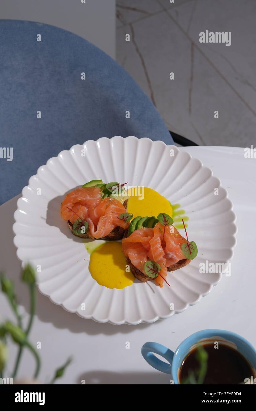 Elegante toast al salmone con salsa cremosa su piatto bianco accanto alla tazza da caffè in un ambiente moderno Foto Stock