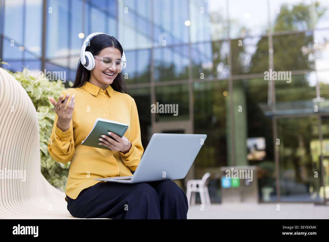 Una giovane donna che fa videoconferenze all'aperto con cuffie e laptop, discute di idee aziendali e prende appunti in uno stile di vita nomade digitale godendo di un lavoro flessibile Foto Stock