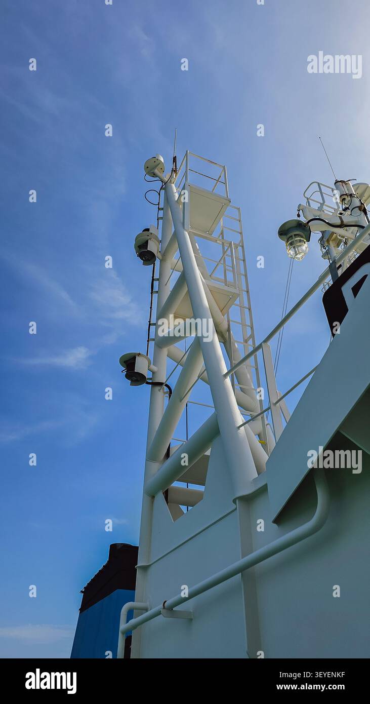 Ripresa verticale ad angolo basso di un albero di nave industriale in metallo bianco con luci di navigazione e scale su una sovrastruttura di un'imbarcazione commerciale sotto una b trasparente Foto Stock