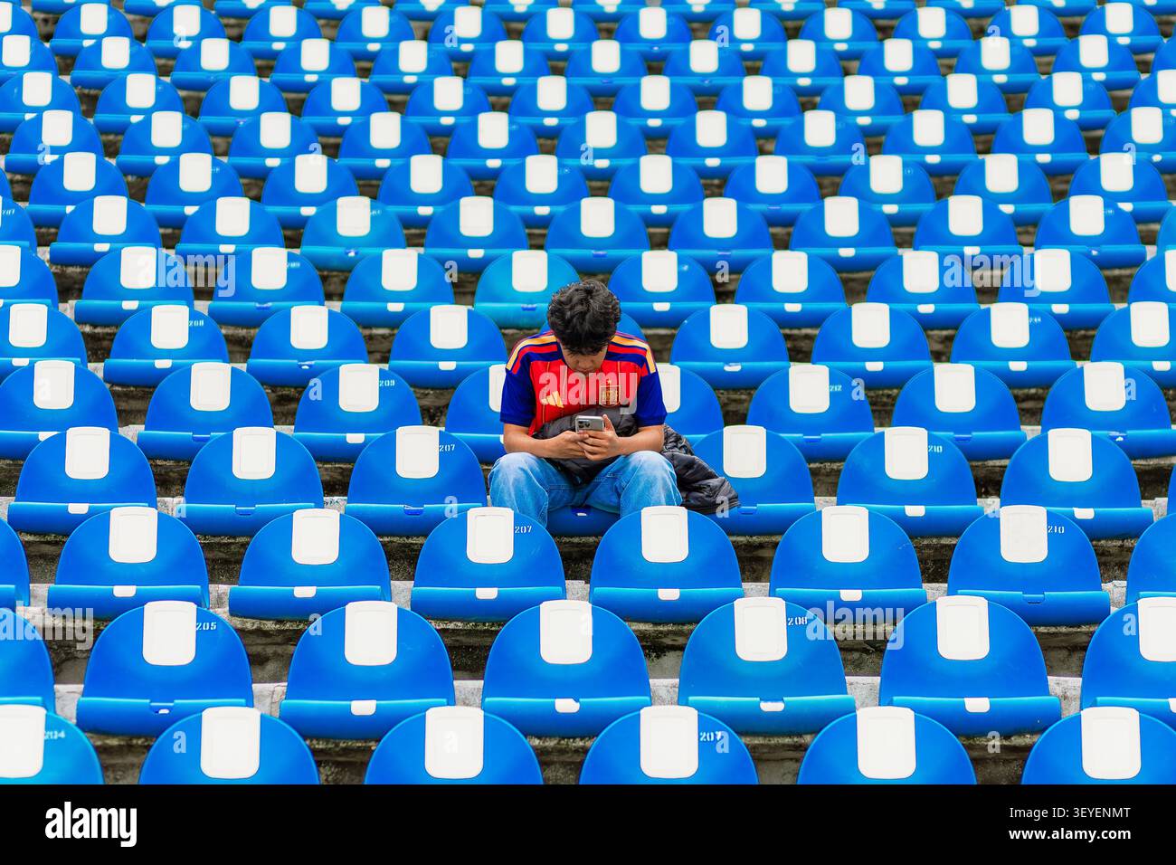 Un singolo spettatore seduto da solo tra file di sedili blu dello stadio guarda il suo smartphone durante il giorno. Foto Stock
