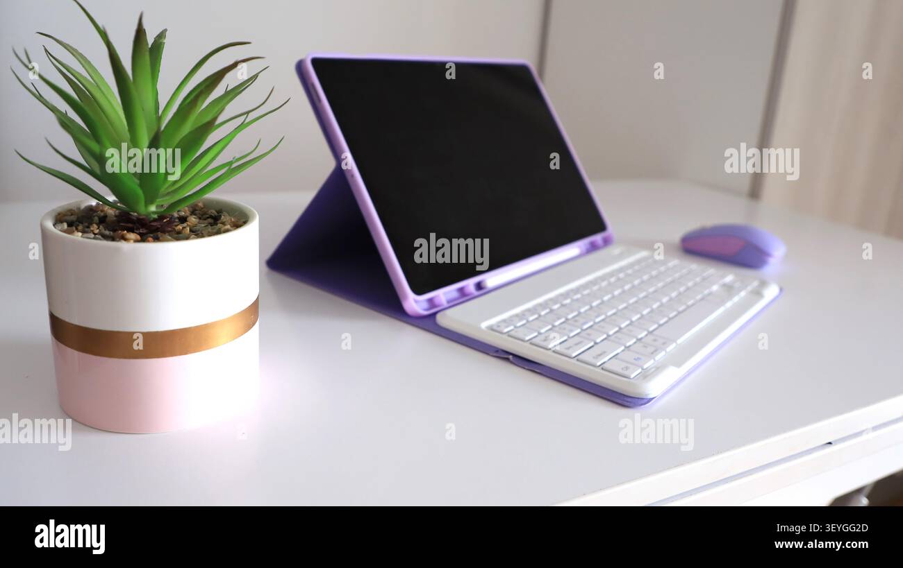 Tablet moderno con custodia viola e mouse wireless su una scrivania bianca accanto a una pianta in vaso decorativa in un ambiente di lavoro contemporaneo Foto Stock