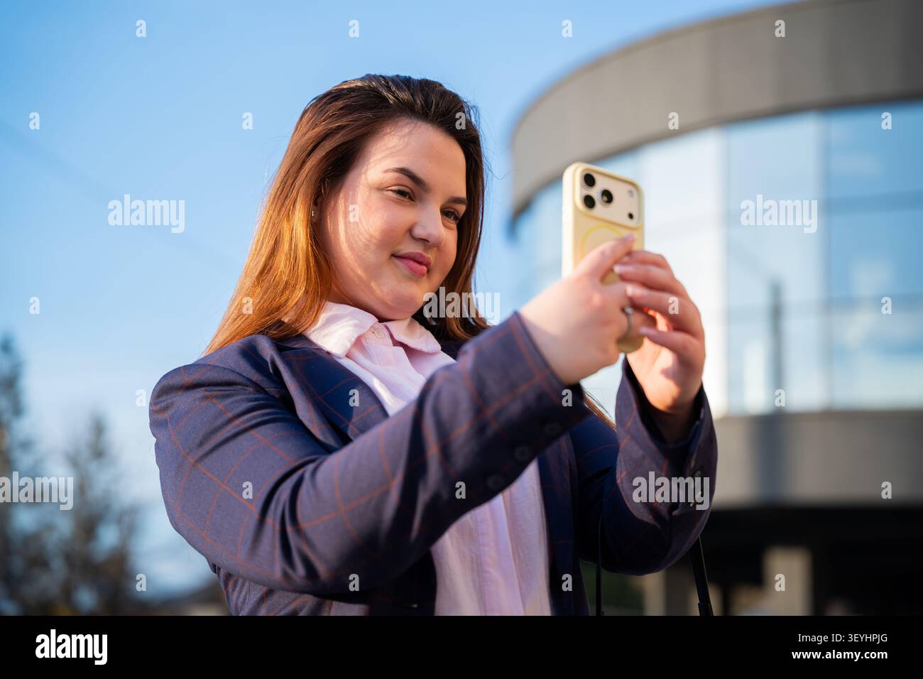 Giovane donna professionale sicura di sé che scatta un selfie con lo smartphone all'esterno di un moderno edificio di vetro. Branding personale comunicazione digitale in Foto Stock