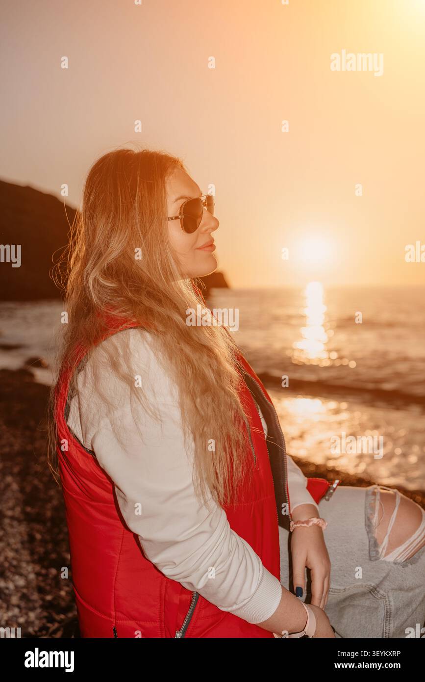 La donna al tramonto guarda le onde dell'oceano al tramonto indossando un gilet rosso e occhiali da sole durante una vacanza tranquilla Foto Stock