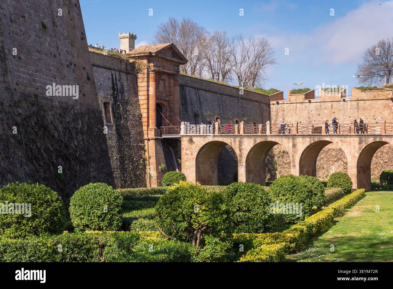 BARCELLONA, SPAGNA - 26 FEBBRAIO 2026: I visitatori camminano su un ponte di pietra accanto alle storiche mura fortificate e ai giardini del castello di Montjuic. Foto Stock