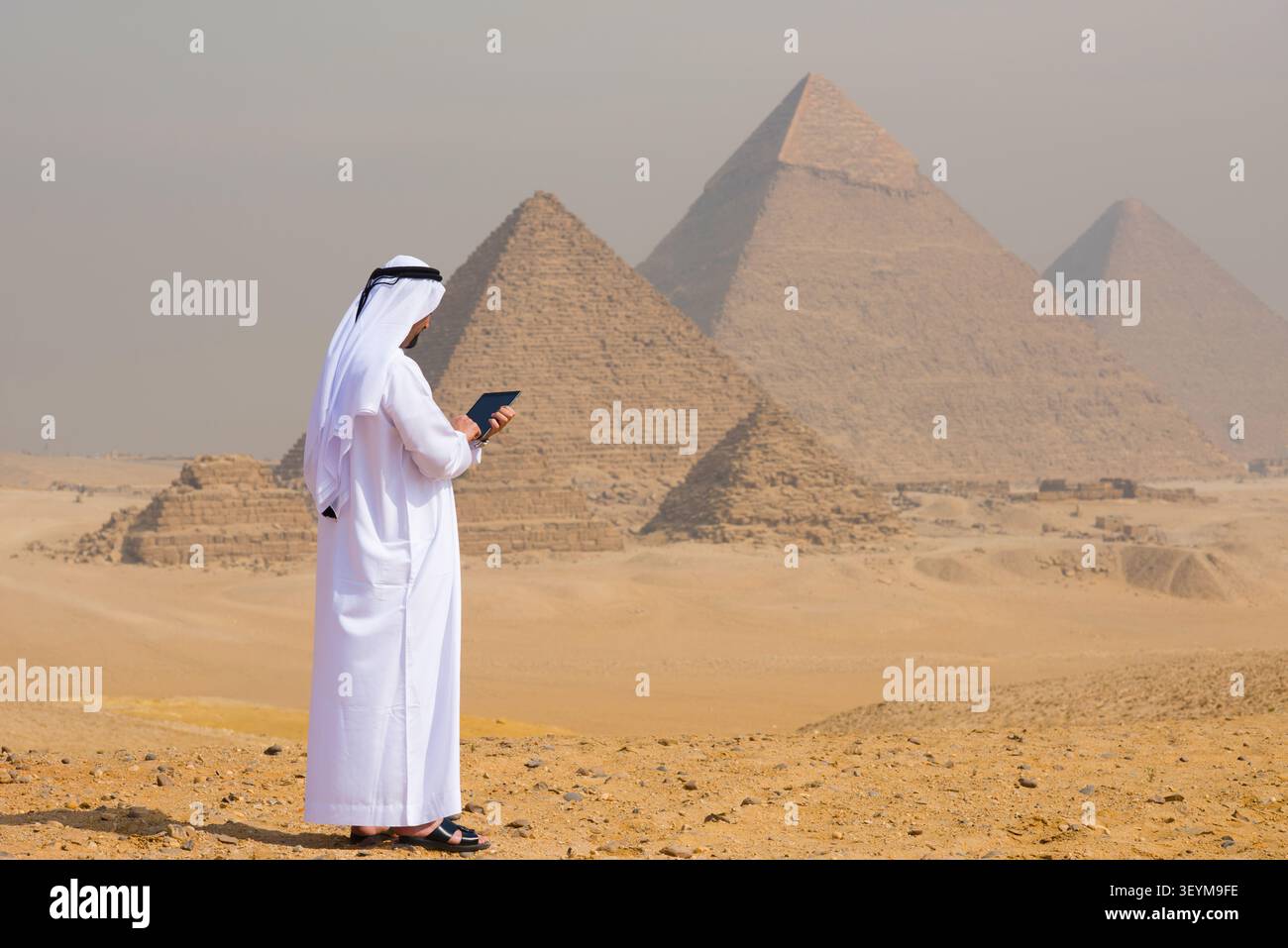 Uomo arabo che indossa un tradizionale abito bianco e keffiyeh utilizzando un tablet digitale mentre si trova sull'altopiano di Giza con le iconiche piramidi che si innalzano nel deserto Foto Stock