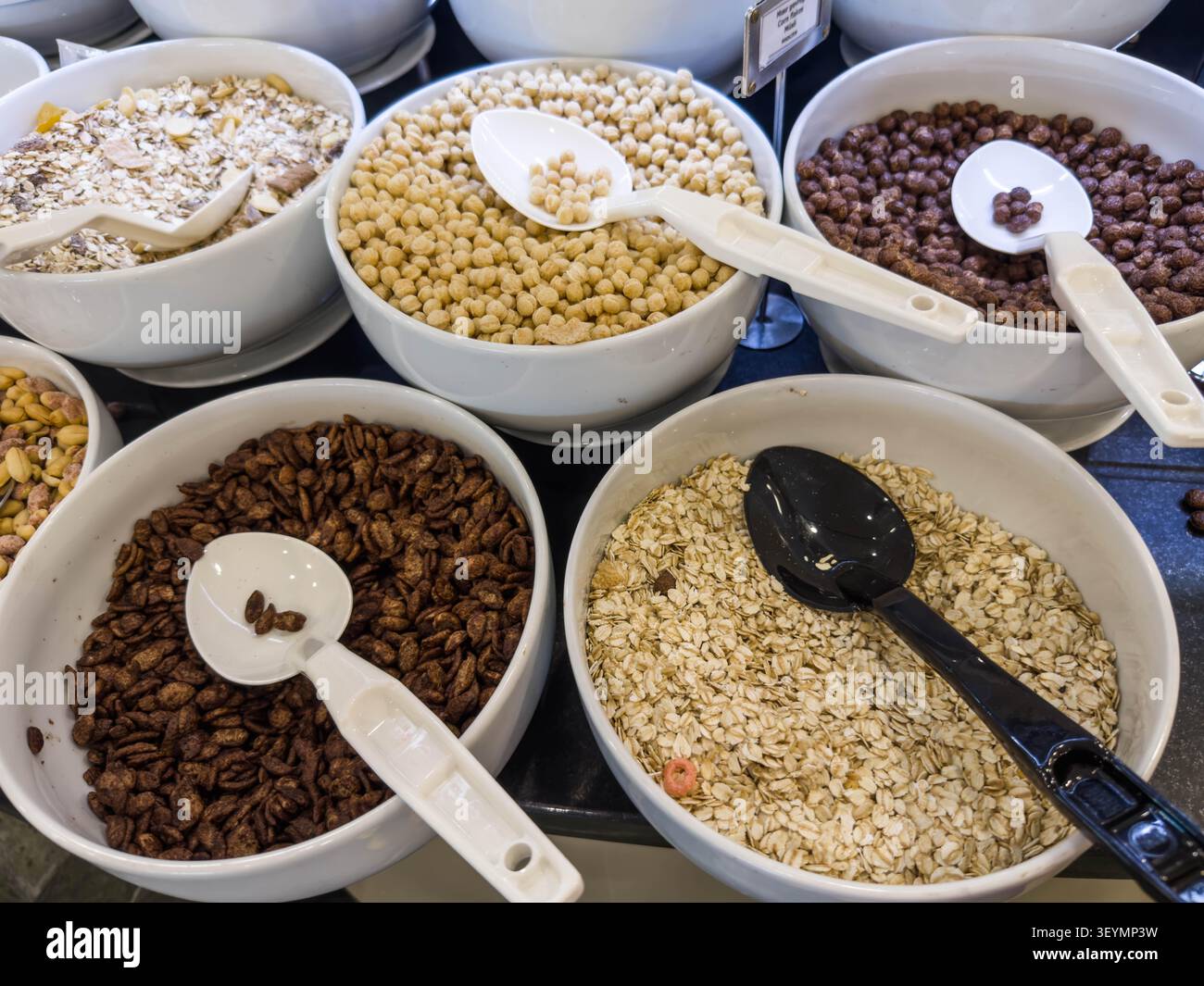 Cereali assortiti per la colazione, grani, noci in ciotole bianche con cucchiai da servire esposti sul tavolo del buffet, scelta di cibi sani scelta nutrizionale concetto di pasto mattutino Foto Stock
