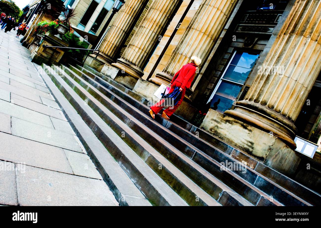 Edimburgo, Scozia, Un’inquadratura a basso angolo, con effetto olandese, di una donna bionda in un vivace completo pantalone rosso che sale i gradini di pietra di un grande edificio neoclassico Foto Stock