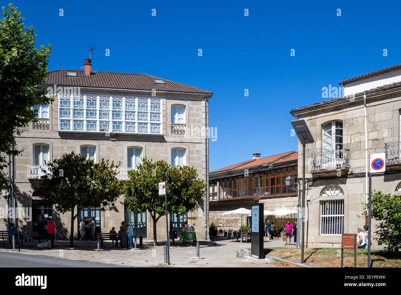 ALLARIZ, SPAGNA - 6 LUGLIO 2025: Edifici tradizionali in granito con una grande galleria di vetro bianca in una piazza pubblica ad Allariz, Ourense, Galizia Foto Stock