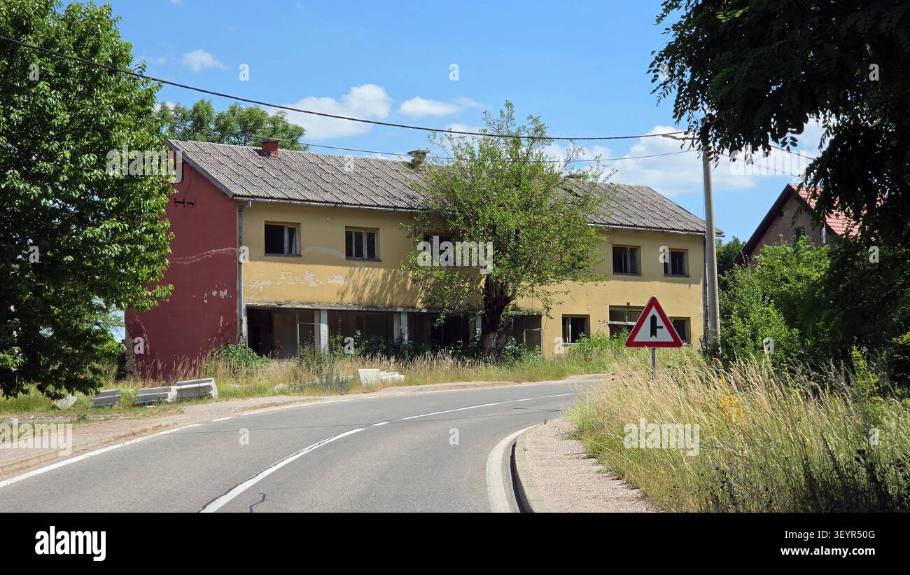 Una struttura residenziale trascurata di colore giallo e rosso si trova su una curva della strada accanto a un segnale stradale triangolare prioritario in una chiara giornata estiva Foto Stock