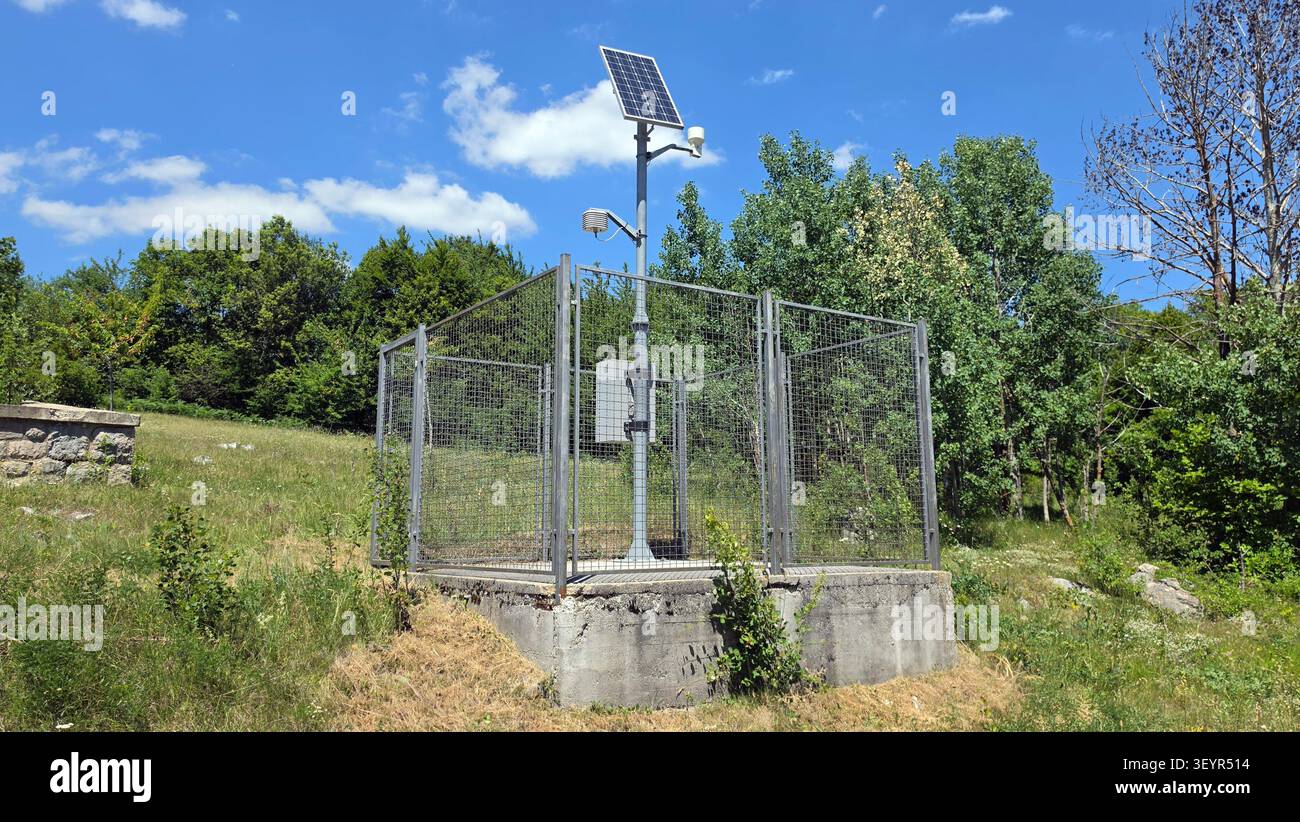 Una stazione meteorologica recintata alimentata da energia solare sorge su una base di cemento in un paesaggio erboso rurale sotto un cielo blu Foto Stock