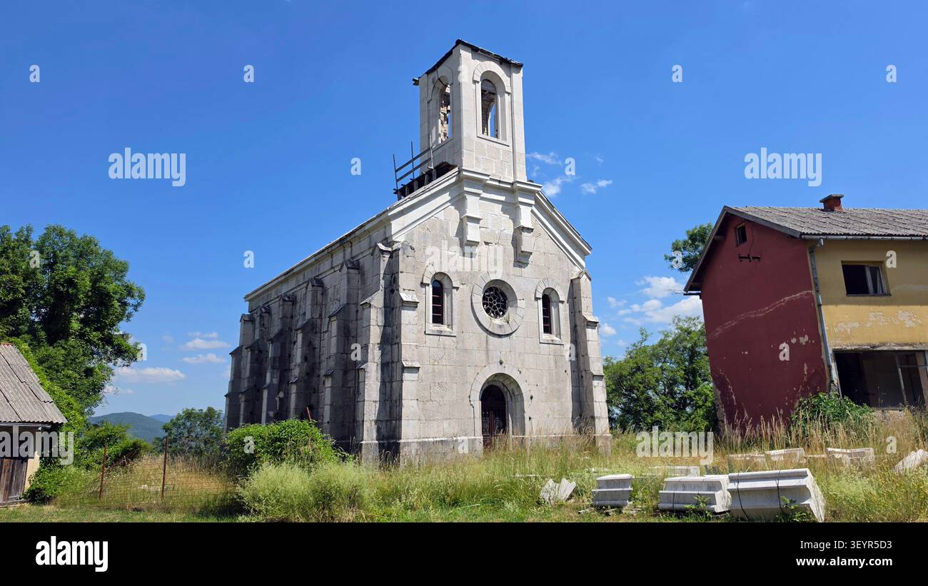 Un'antica chiesa in pietra con finestre ad arco e dettagliata facciata in muratura sorge in un cortile erboso sovrastato accanto ad una casa a due piani sotto un cielo azzurro Foto Stock