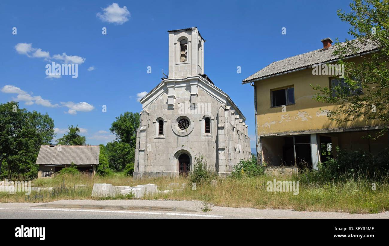 Una storica chiesa in pietra concava con un ingresso ad arco si trova accanto a un edificio giallo in decadenza tra erba selvatica sotto un cielo estivo limpido Foto Stock