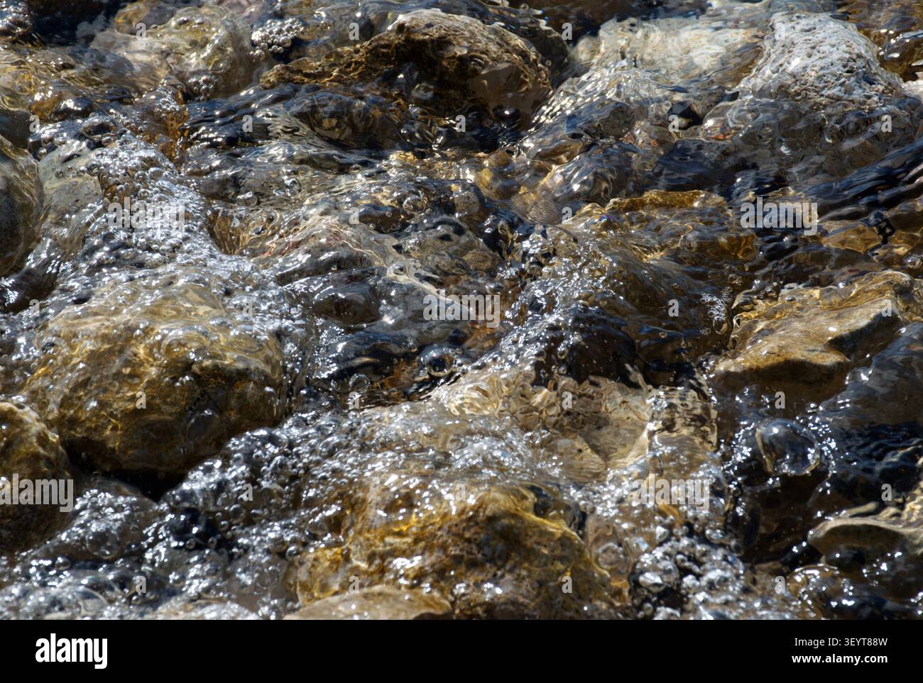 Primo piano di acqua di mare limpida che scorre su rocce naturali lungo una costa rocciosa mediterranea. La luce del sole evidenzia le pietre strutturate e le onde gentili Foto Stock