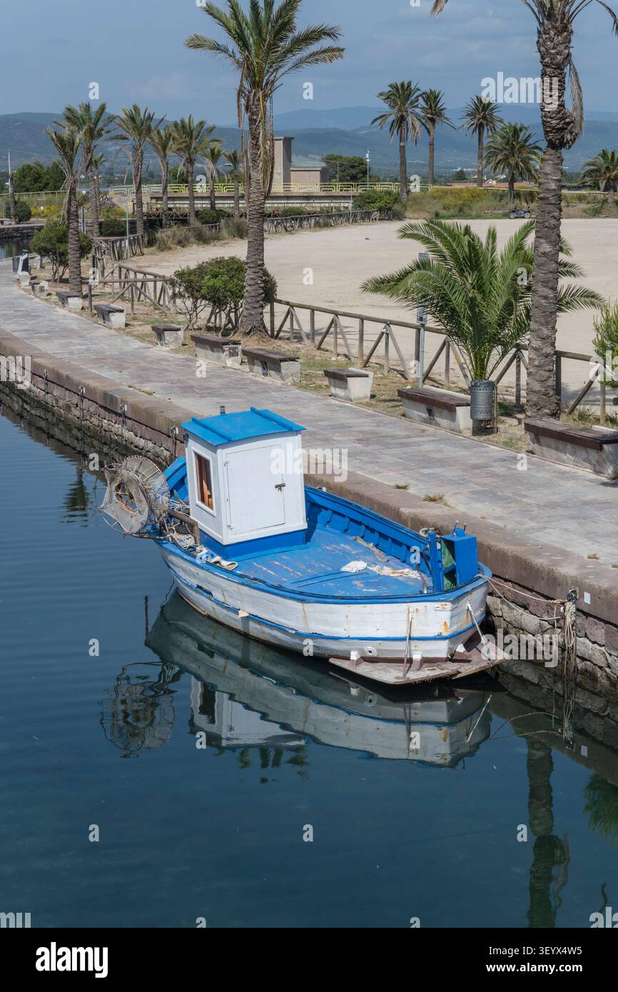 Paesaggio urbano con una piccola barca da pesca sull'argine del porto dei canali sulla costa mediterranea, sparato alla luce della primavera a Porto Pino, Cagliari, Italia Foto Stock
